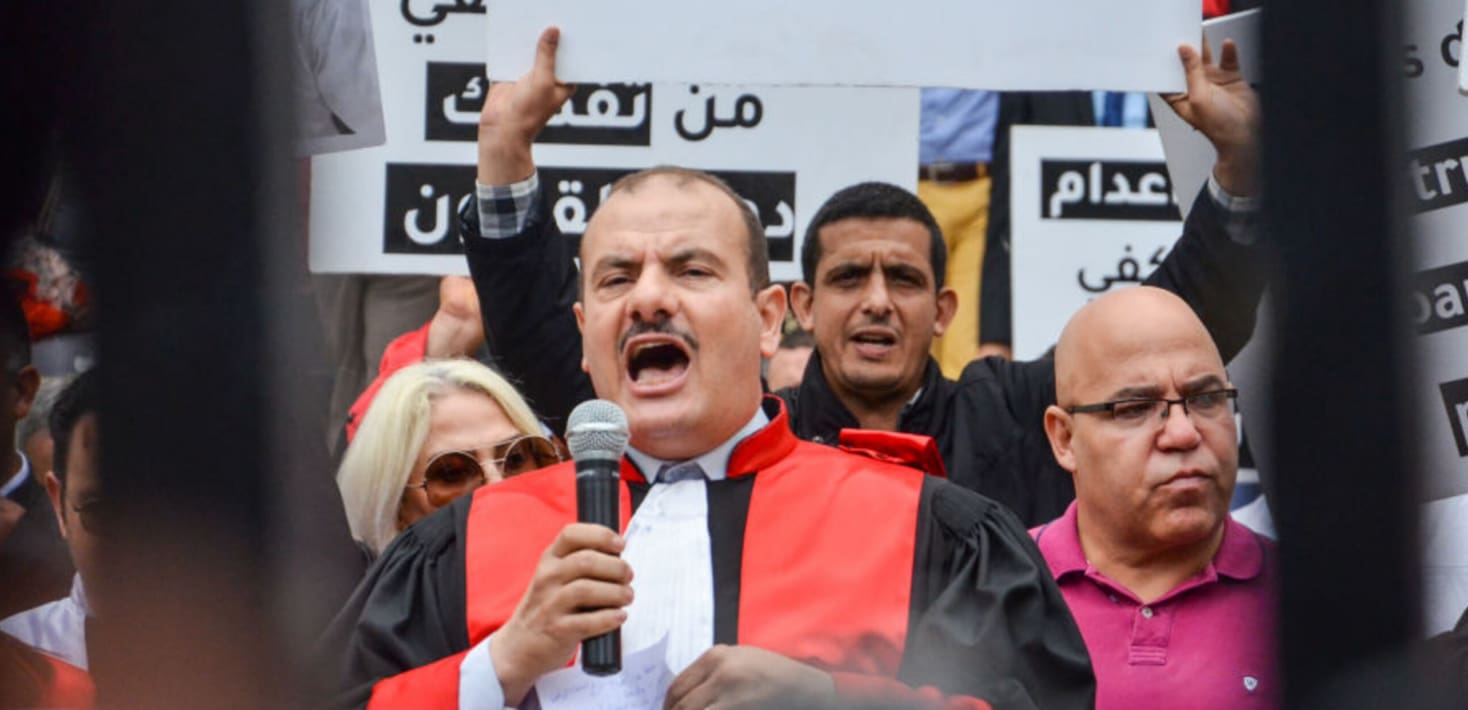 Middle aged bald man with a mustache, speaking with a microphone and wearing a judge's robes. He is orating to a crowd of people around him.