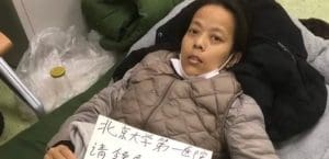 A middle-age Chinese woman lying in a hospital bed holding a sign written in Mandarin or Cantonese.