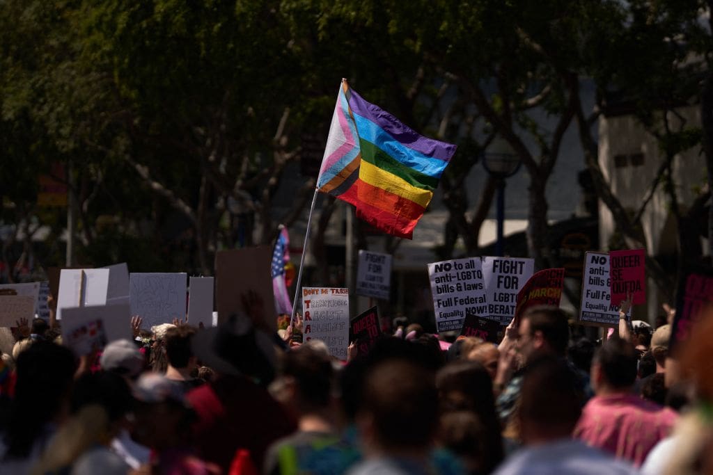 An LGBTQ+ flag over a protest crowd for LGBTQ+ rights.