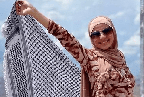 A smiling young woman in a tan hijab, holding up a keffiyeh.