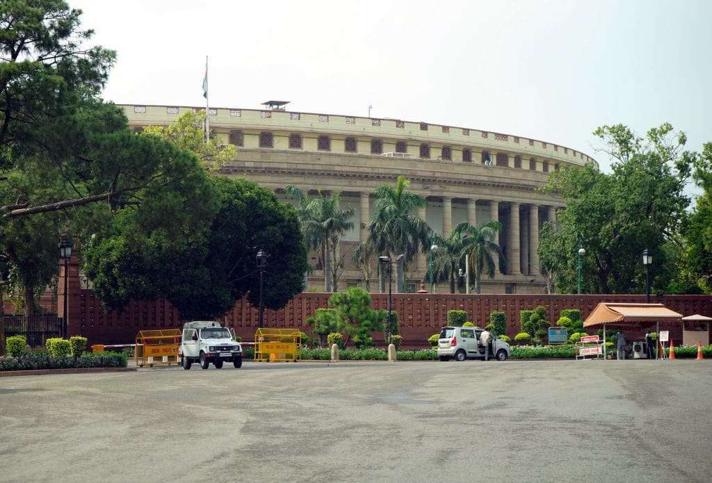The Lok Sabha building (Lower House of India’s Parliament). Circular, beige building with Romanesque columns surrounded by palm trees and a red gate.