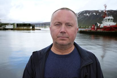 A middle-aged, bald Caucasian man wearing a black jacket and navy blue shirt. He is standing in front of a body of water with a boat in the background.