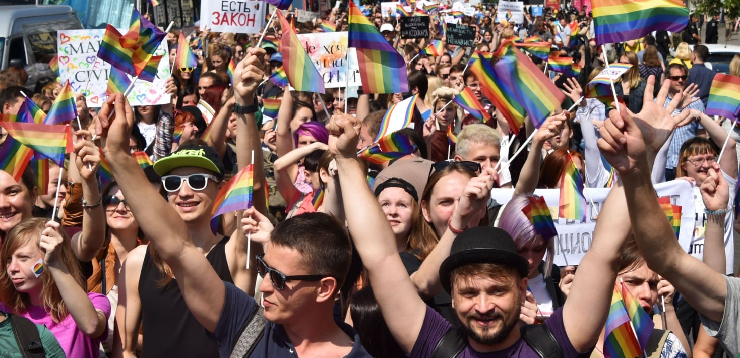 A large crowd of people cheering and holding mini LGBTQ+ flags in the air. Many are also holding up signs written in Ukrainian.