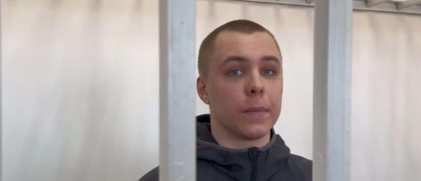A young Caucasian male behind steel bars. He is looking directly at the camera, no smile.