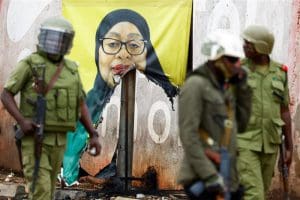 Armed soldiers in green uniforms and riot helmets stand in front of a torn banner of Tanzanian President Samia Suluhu.