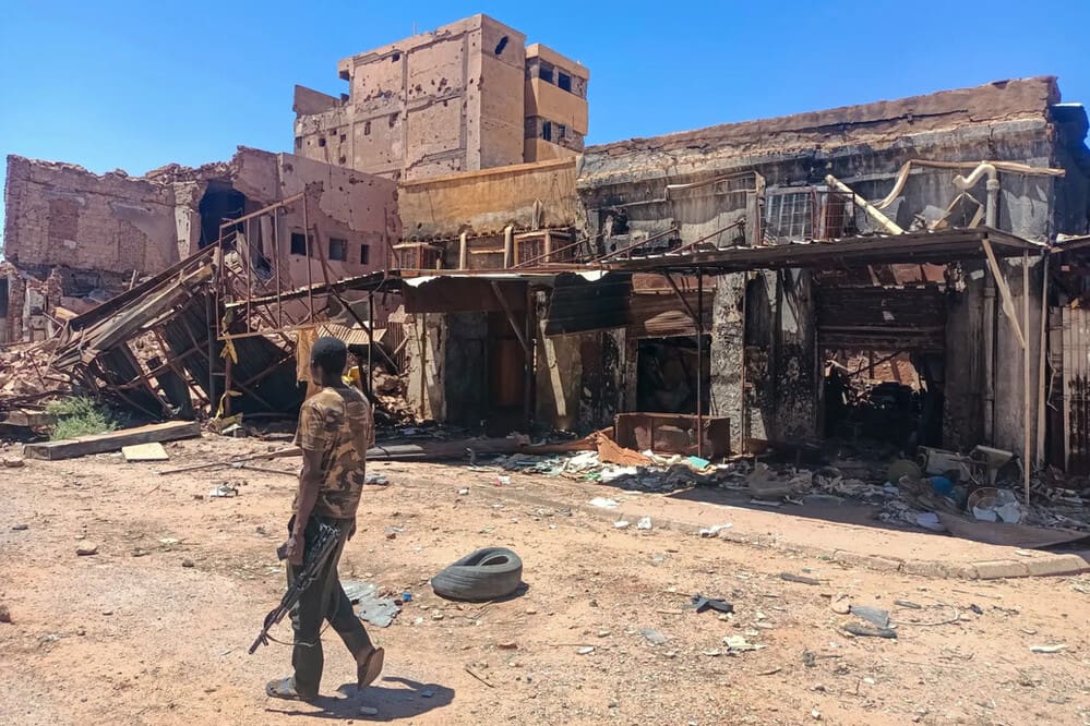 A soldier walks down a deserted street, lined with ruined burned out shops.