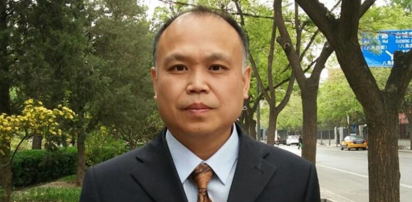 A photograph of a Chinese man wearing a blue collared shirt, orange tie and black blazer jacket.