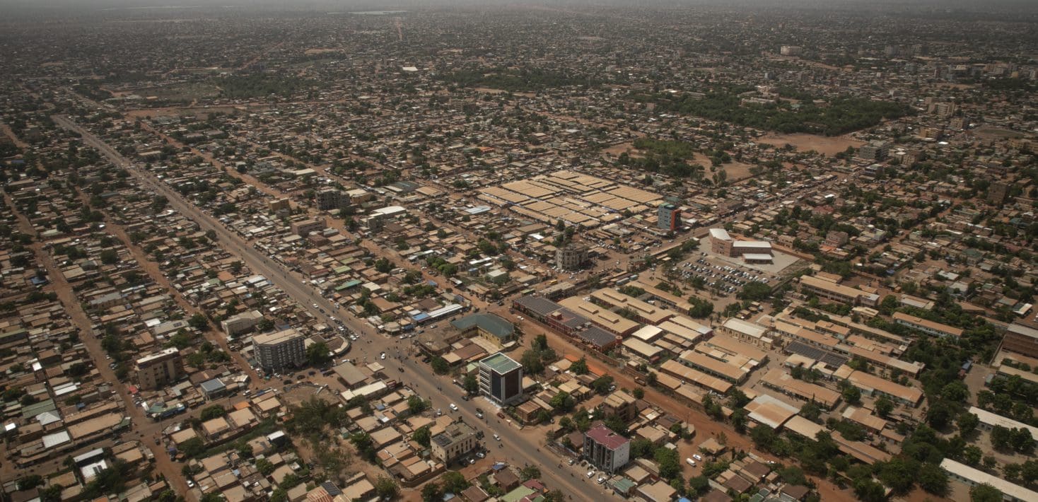 An aerial view of Niamey, the capital of Niger, a grid of tan and brown buildings with some paved roads dotted with low trees. It looks hot.