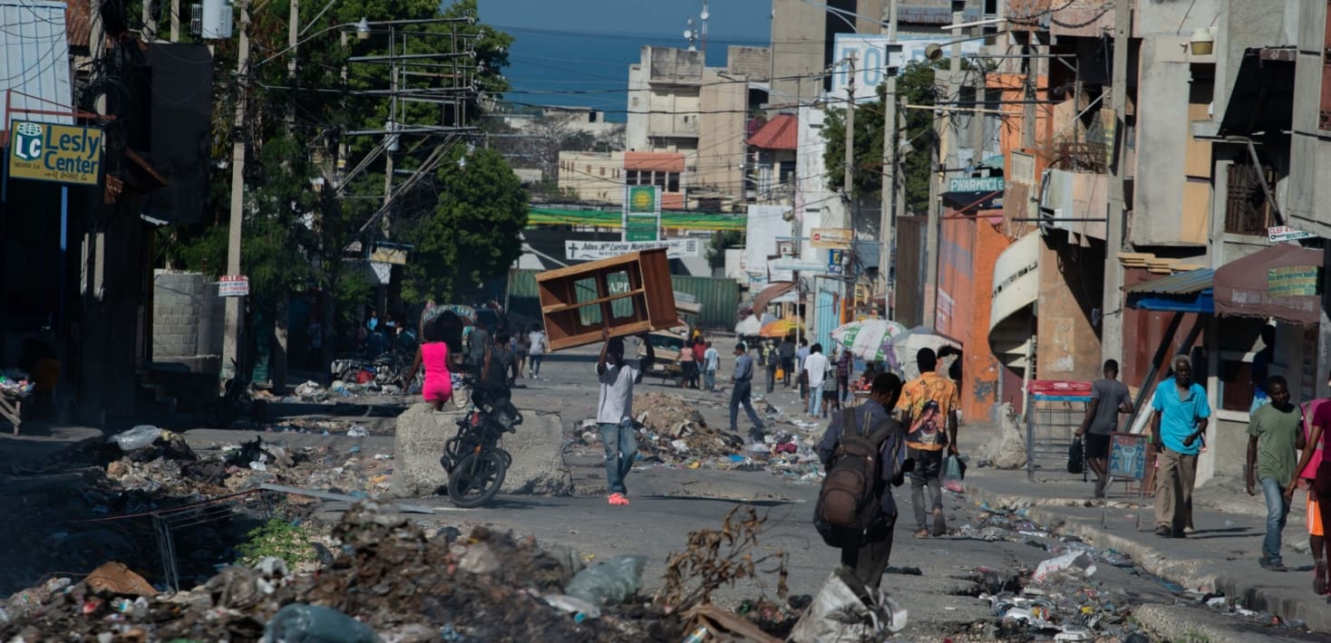 A street lined with shops, people walking on the sidewalks, and rubble, trash and broken furniture surrounding the street.