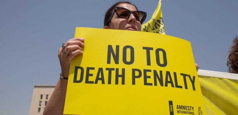 A woman wearing sunglasses holding a yellow sign that says "No To Death Penalty"