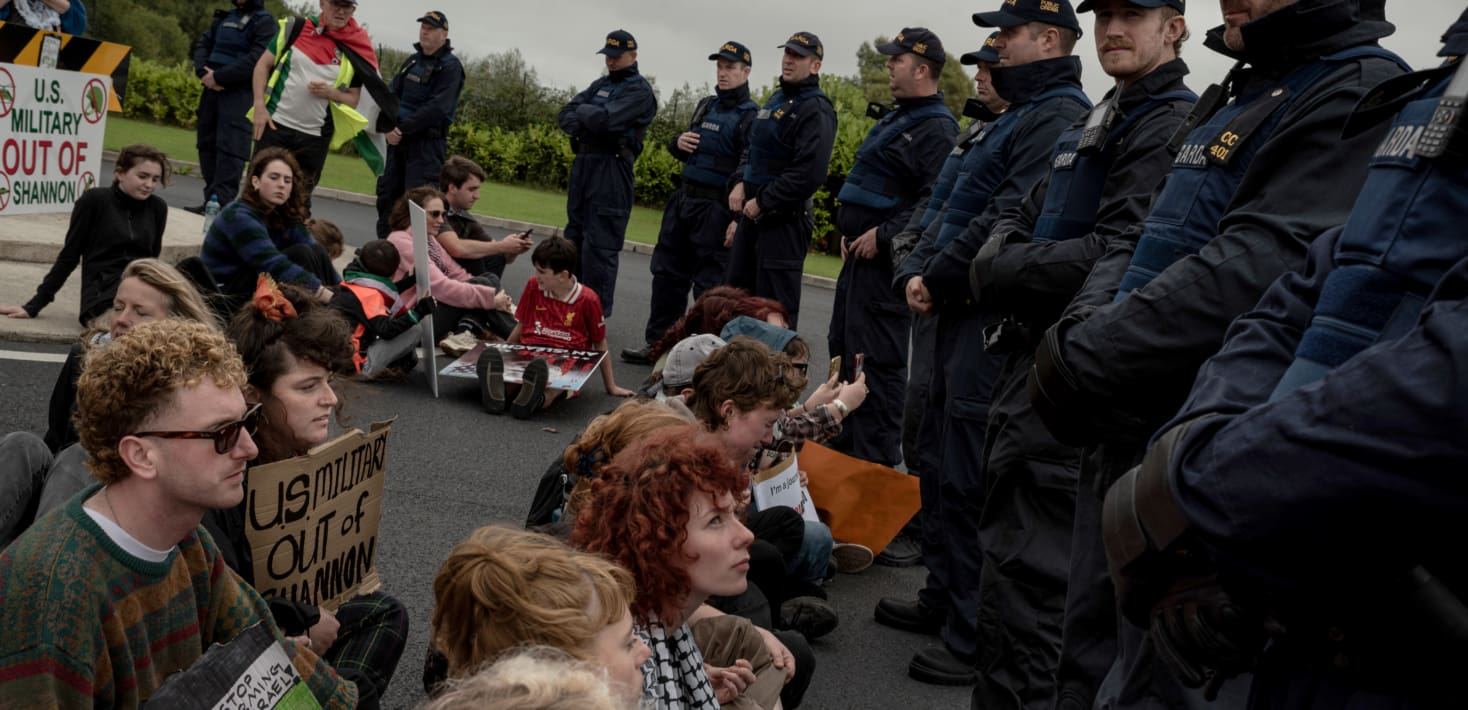 A group of largely young people sitting on the street with protest signs, surrounded by police officers. The signs say 'U.S. military out of Shannon'.