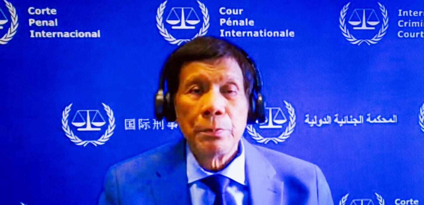 Former Philippines President Rodrigo Duterte sits in the dock at a International Criminal Court wearing a blue suit and tie and headphones, in front of a blue ICC branded background.