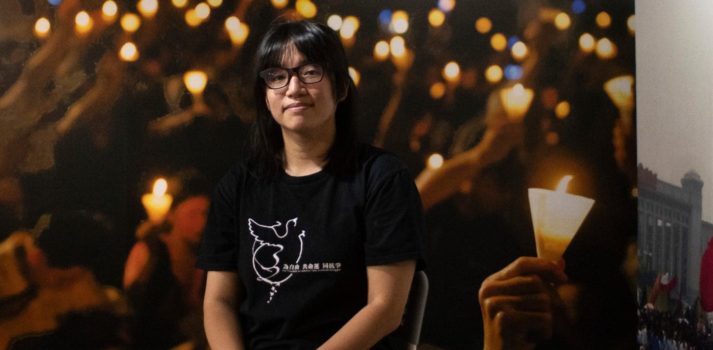 Chow Hang-tung, wearing a black tshirt, stands in front of a backdrop showing hands holding vigil candles.