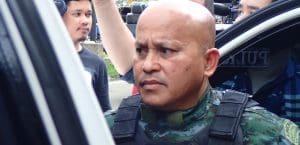 Ronald “Bato” dela Rosa, with a shaved head and wearing a thick police uniform, stands behind the open door to a car, looking to his right, with TV journalists behind him.