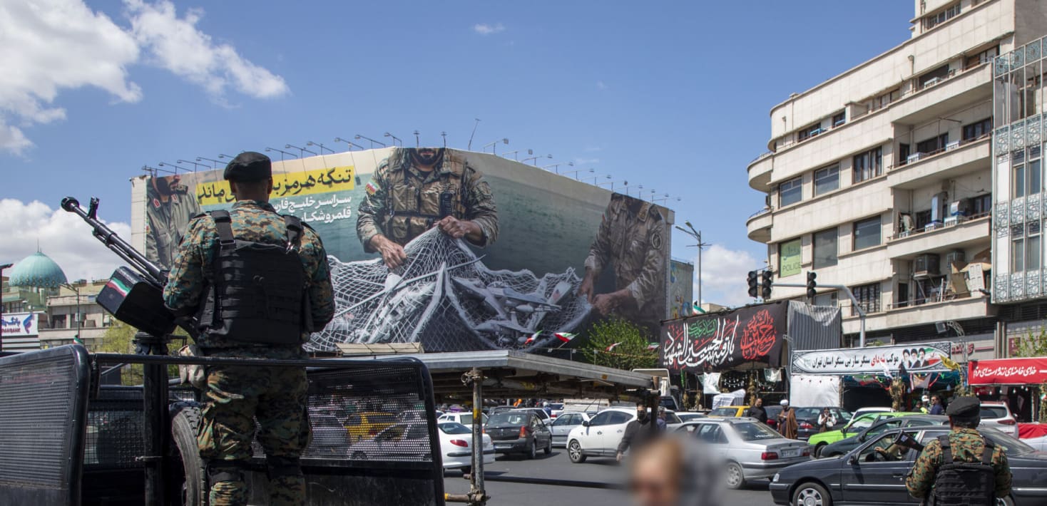 A uniformed soldier stands behind a heavy machine gun mounted on the back of a truck, overseeing a busy city street beneath a large billboard showing Iranian military figures capturing western fighter jets with a net.
