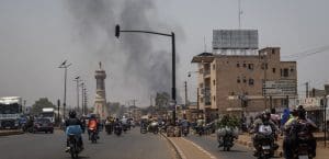 Smoke rises on the horizon behind a cityscape of a road and low buildings with people driving motorcycles.