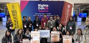 A group of people poses for a picture, most of them smiling, in front of a yellow Amnesty International banner with Chinese writing, and a blue RightsCon25 Taipei backdrop. They are holding signs about human rights and open culture.