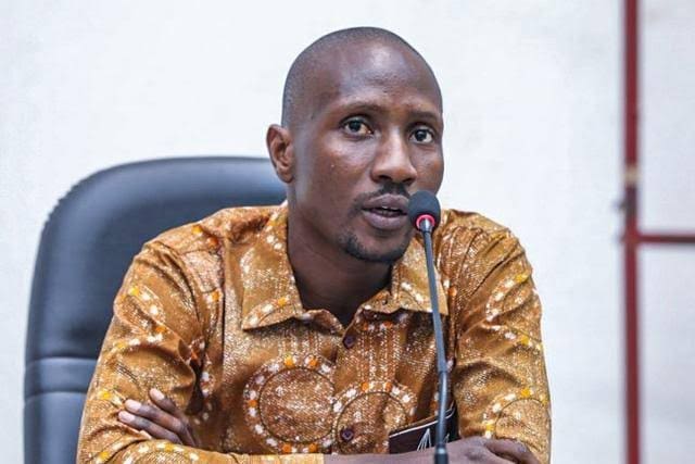 Journalist Atiana Serge Oulon wearing a colorful shirt, sits at an office chair and speaks into a microphone. He is leaning forward slightly with a calm and thoughtful expression on his face. The room has white walls.