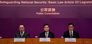 Hong Kong Chief Executive John Lee, in a dark suit and white tie, sits in the center of a panel between Secretary for Security Tang Ping-keung and Secretary for Security Paul Lam, beneath a banner reading: 