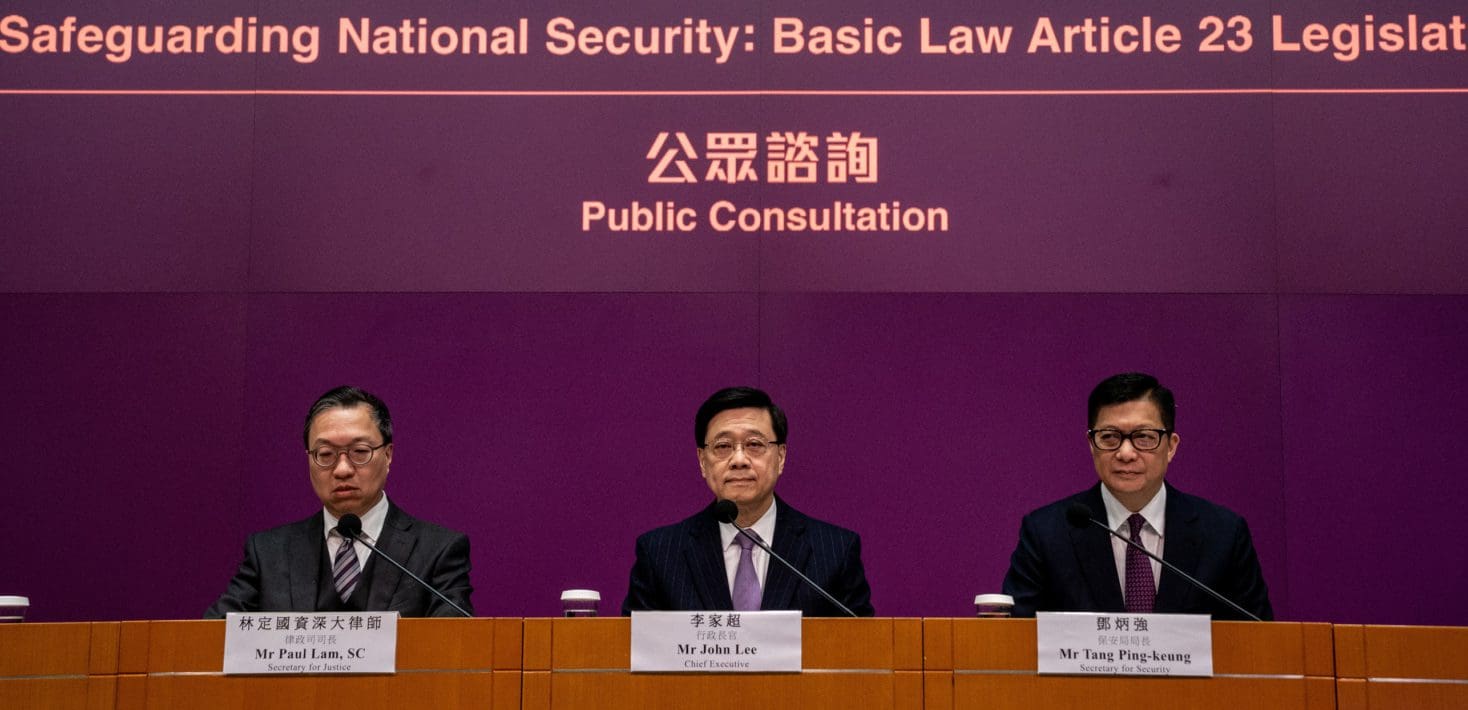 Hong Kong Chief Executive John Lee, in a dark suit and white tie, sits in the center of a panel between Secretary for Security Tang Ping-keung and Secretary for Security Paul Lam, beneath a banner reading: 