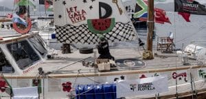 A collection of sailboats at harbor with flags and painted signs showing solidarity with Gaza.