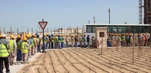 A line of workers entering a secured compound under a dusty sky in what appears to be hot weather.