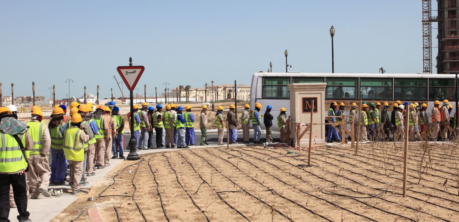 A line of workers entering a secured compound under a dusty sky in what appears to be hot weather.