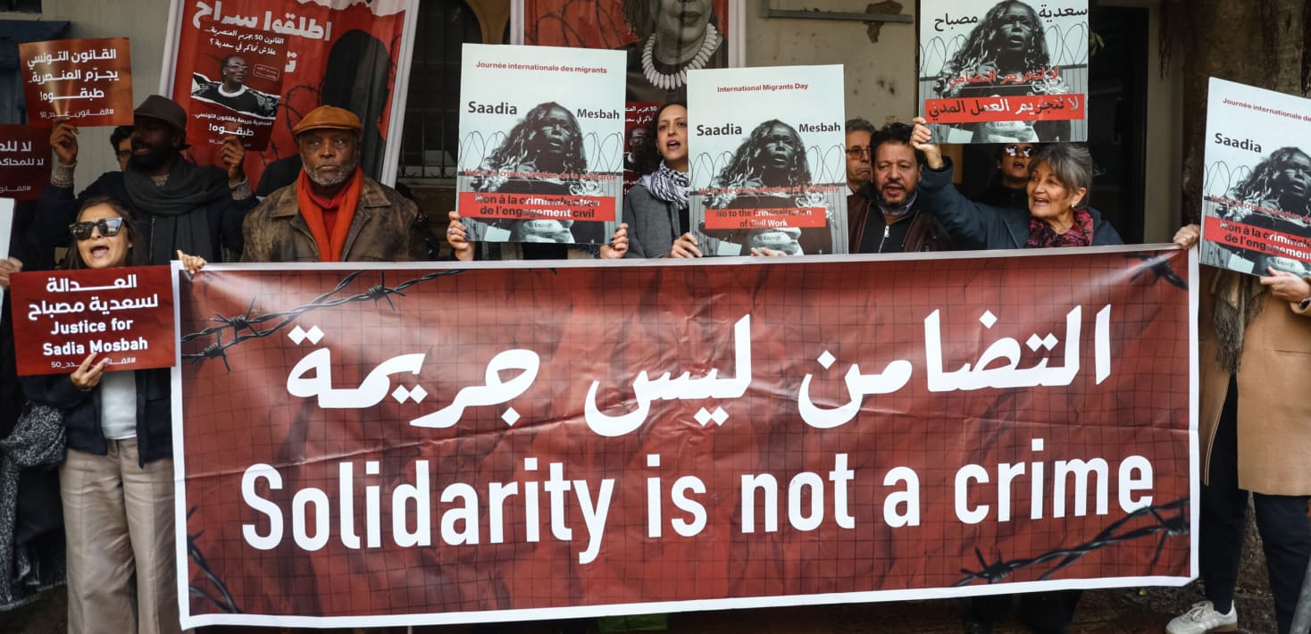 A group of racially diverse protesters hold signs with the face of Saadia Mosbah speaking in front of barbed wire, and hold a large red banner with white letters in English and Arabic reading 