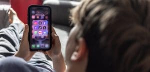 A teenagen boy with blond hair lounges on a couch, looking at a phone screen with the logos of social media apps like facebook, tiktok, twitter, whatsapp, etc.