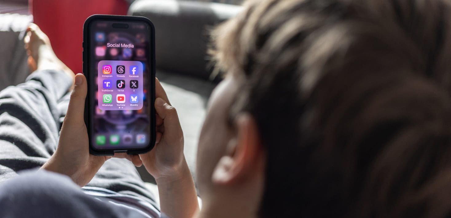 A teenagen boy with blond hair lounges on a couch, looking at a phone screen with the logos of social media apps like facebook, tiktok, twitter, whatsapp, etc.