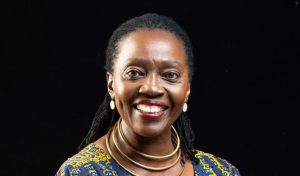 Martha Karua smiles as she poses for a portrait wearing a colorful outfit against a black background.