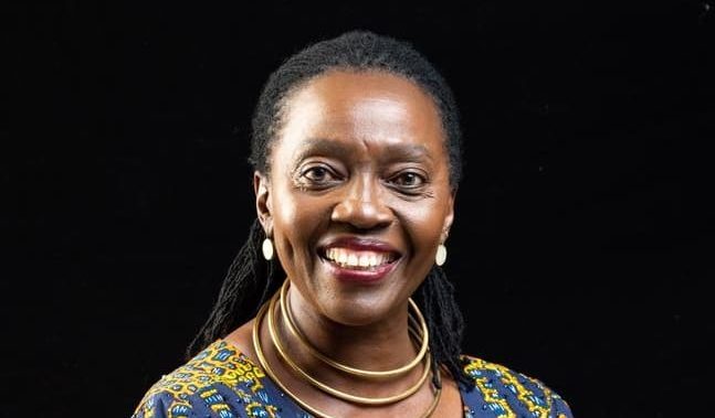 Martha Karua smiles as she poses for a portrait wearing a colorful outfit against a black background.