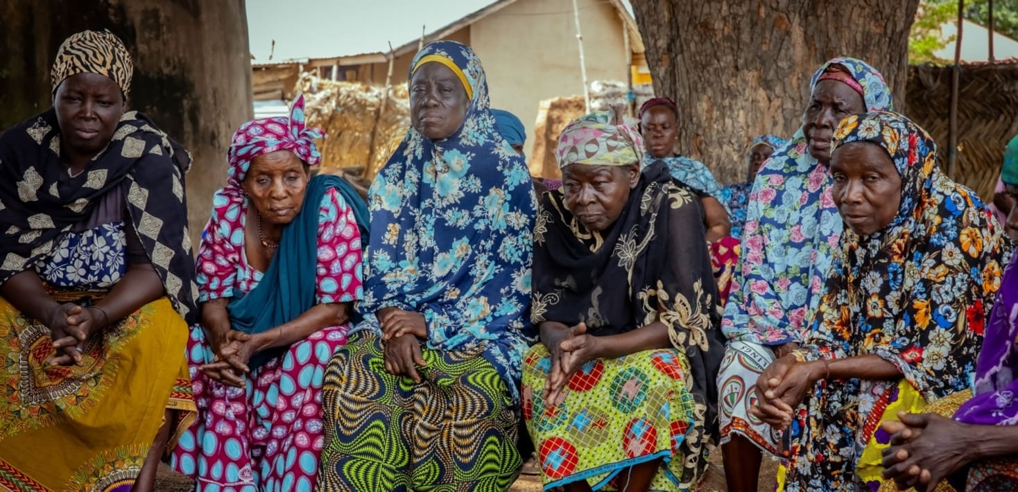 Six older women in brightly colored clothing, sit together under shade trees, despondent expressions on their faces.
