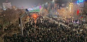 A crowd of people, mostly wearing black, fills a street in a dark, foggy night. Two men climb what appears to be a scaffold or a light pole that hangs over the street. A fire burns.
