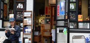 A man wearing a baseball cap and glasses reads a book in front of a bookshop.