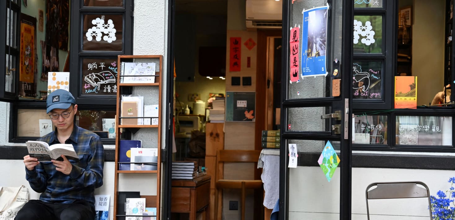 A man wearing a baseball cap and glasses reads a book in front of a bookshop.