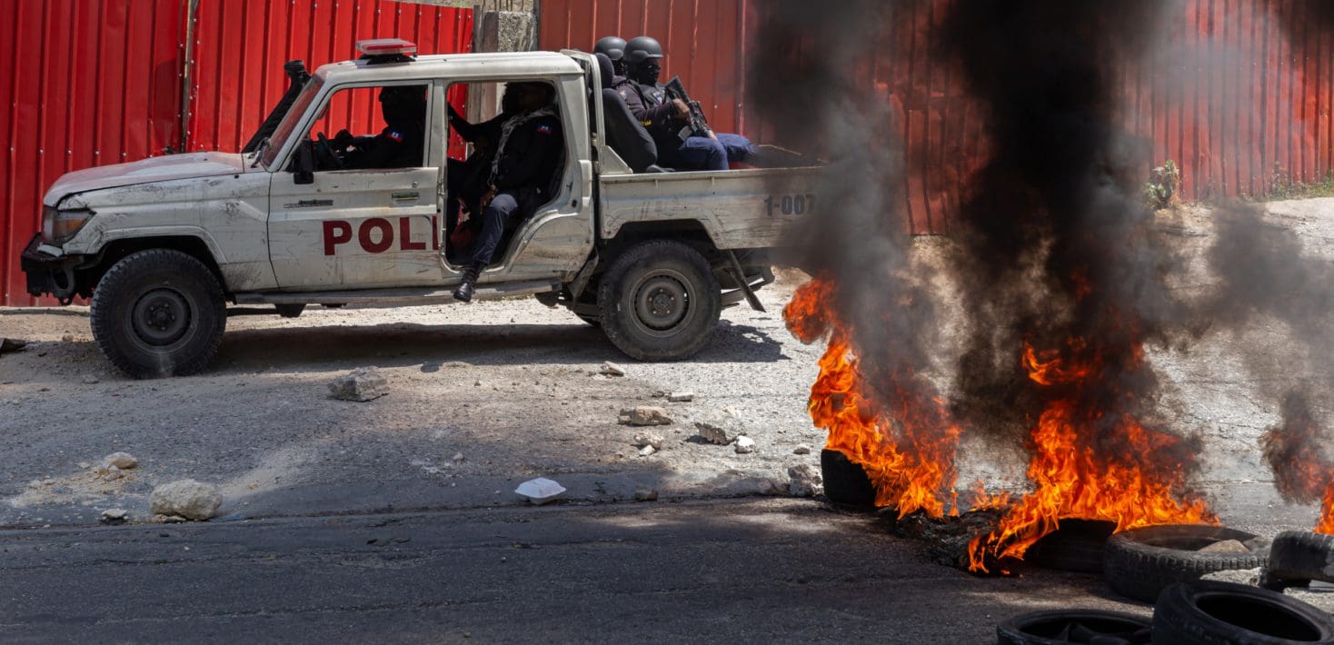 A white pick up truck with POLICE written on the side loaded with armed men in uniform and riot gear pulls up alongside some burning tires.