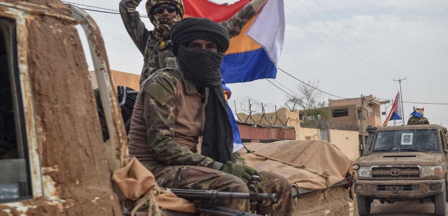 A soldier in combat fatigues and a black headscarf sits in the back of a military pick up, looking blankly at the camera, a machine gun barrel poking out beside him. Behind him, another soldier wearing sunglasses waves a red, white, blue, and green flag.