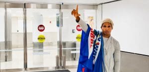 Luis Manuel Otero Alcántara wears a knit cap, gray hoodie, and a Cuban flag over his shoulder, while holding his right arm in the air with his pointer finger and thumb outstretched, in front of glass 