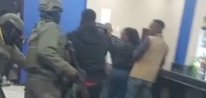 A blurry image shows armored police holding AK-47s holding another man in custody in what appears to be an office or inside a building with other bystanders watching.
