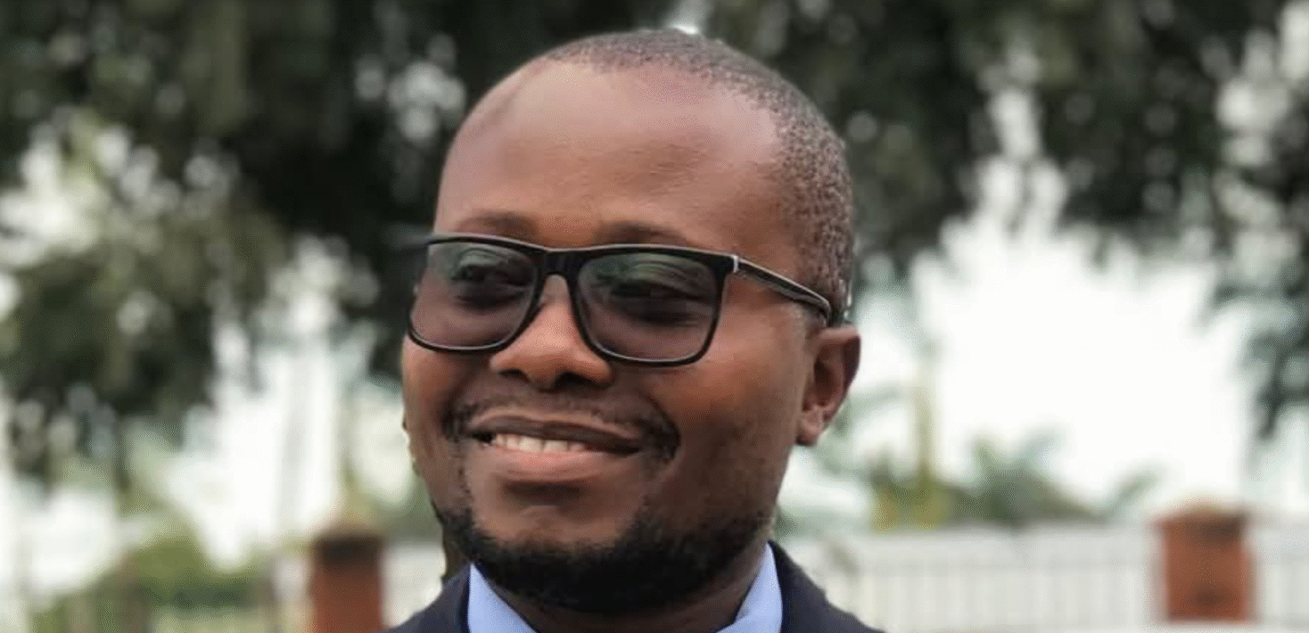 MacPherson Mukulka, wearing black rimmed glasses and a suit, smiles and looks slightly off camera.