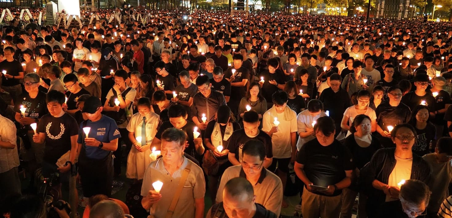 A crowd of thousands of people, each holding a candle with their head bowed in silence, fills a public square at night.