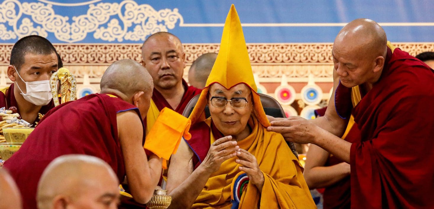 The Dalai Lama, wearing glasses, a slight smile, red and orange robes, and a pointed orange cap, is surrounded by monks in similarly colored robes, in front of an ornate blue, red, and gold tapestry.