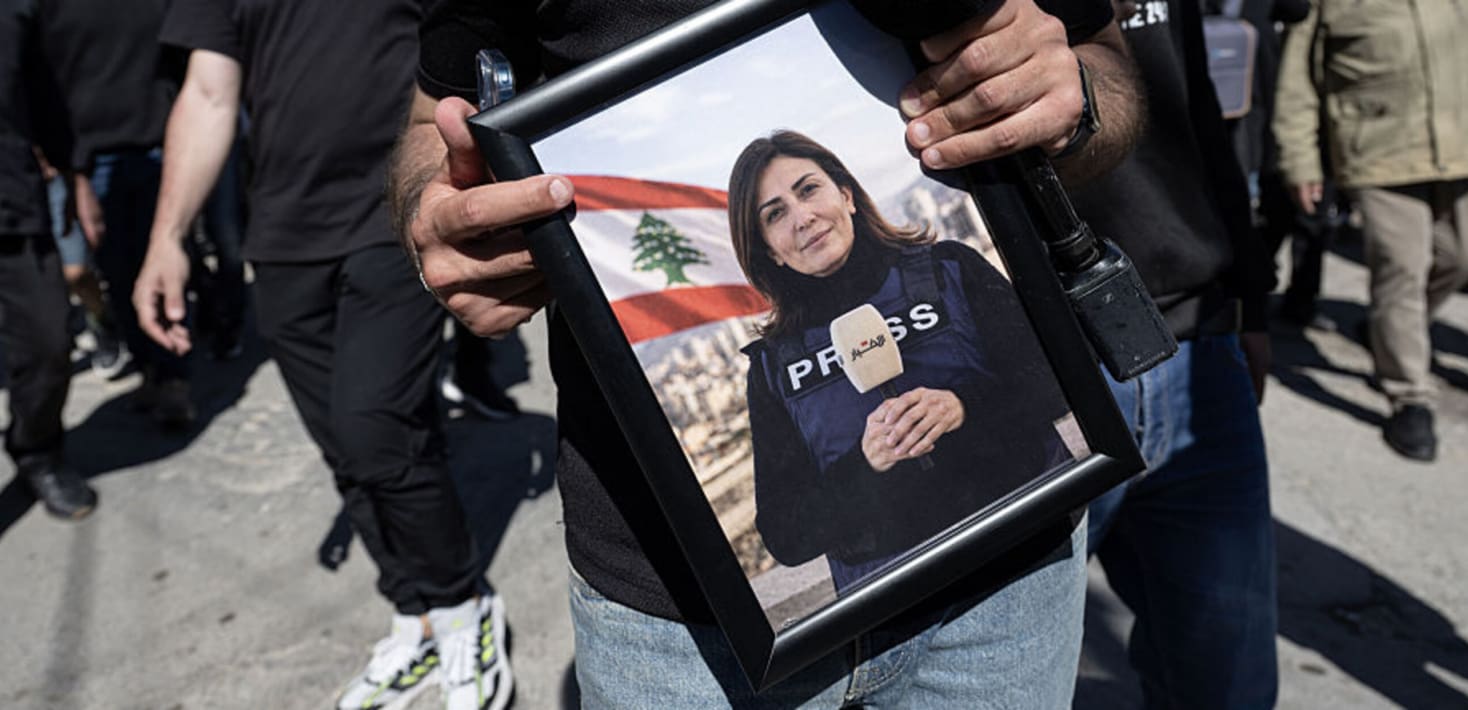 A person with a camera slung over their shoulder holds a framed photograph of Amal Khalil holding a micrphone and wearing a press vest in front of the Lebanese flag.