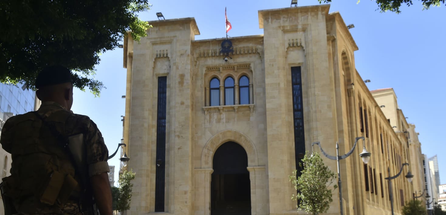 A tall cathedral-looking building with a Lebanese flag on top. A soldier looks up at it.