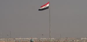 The Iraqi flag flies on an enormously tall flagpole above a barely visible cityscape of low, nondescript, cream colored buildings and electric wires against a sky gray with dust.