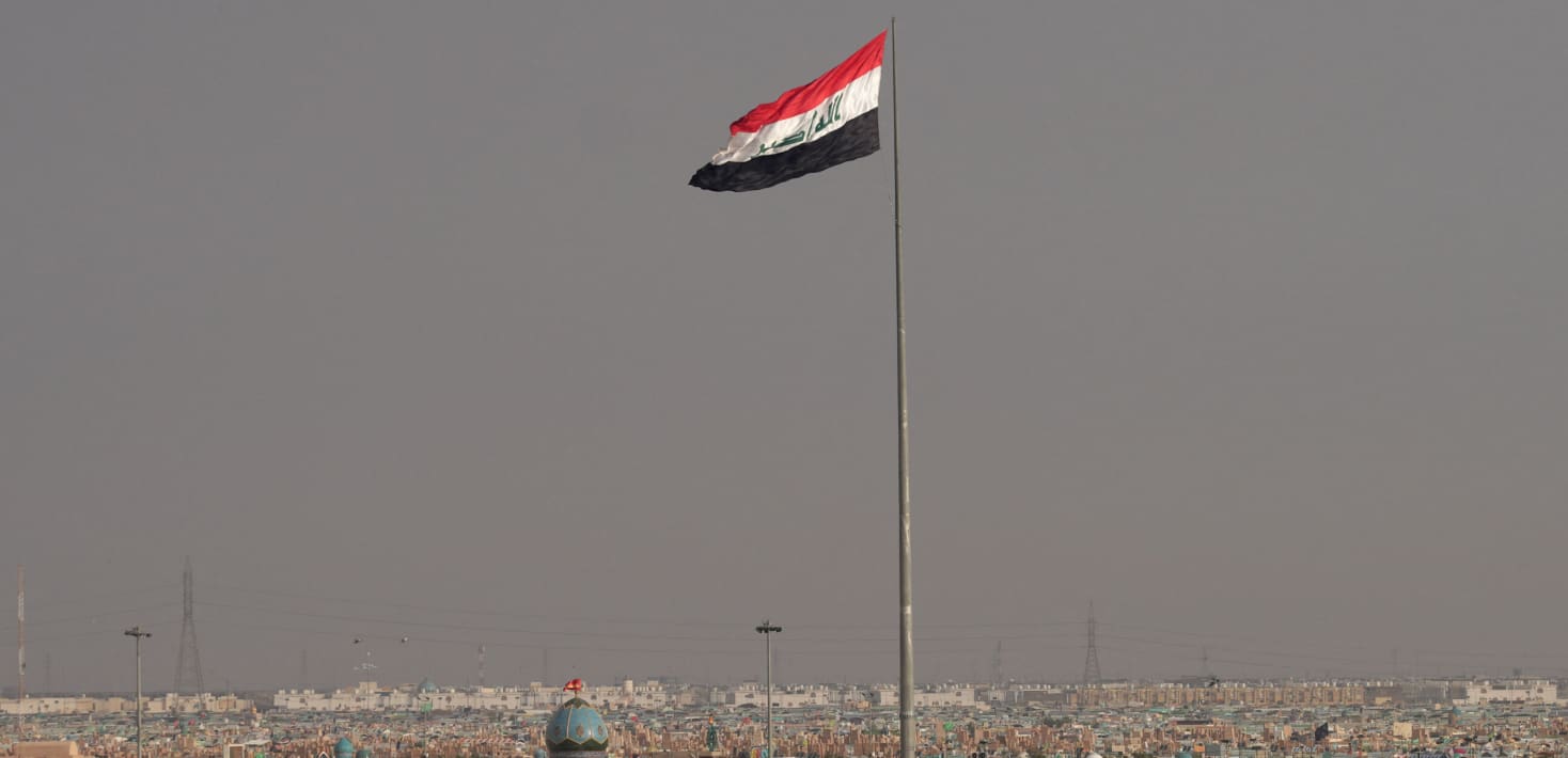 The Iraqi flag flies on an enormously tall flagpole above a barely visible cityscape of low, nondescript, cream colored buildings and electric wires against a sky gray with dust.