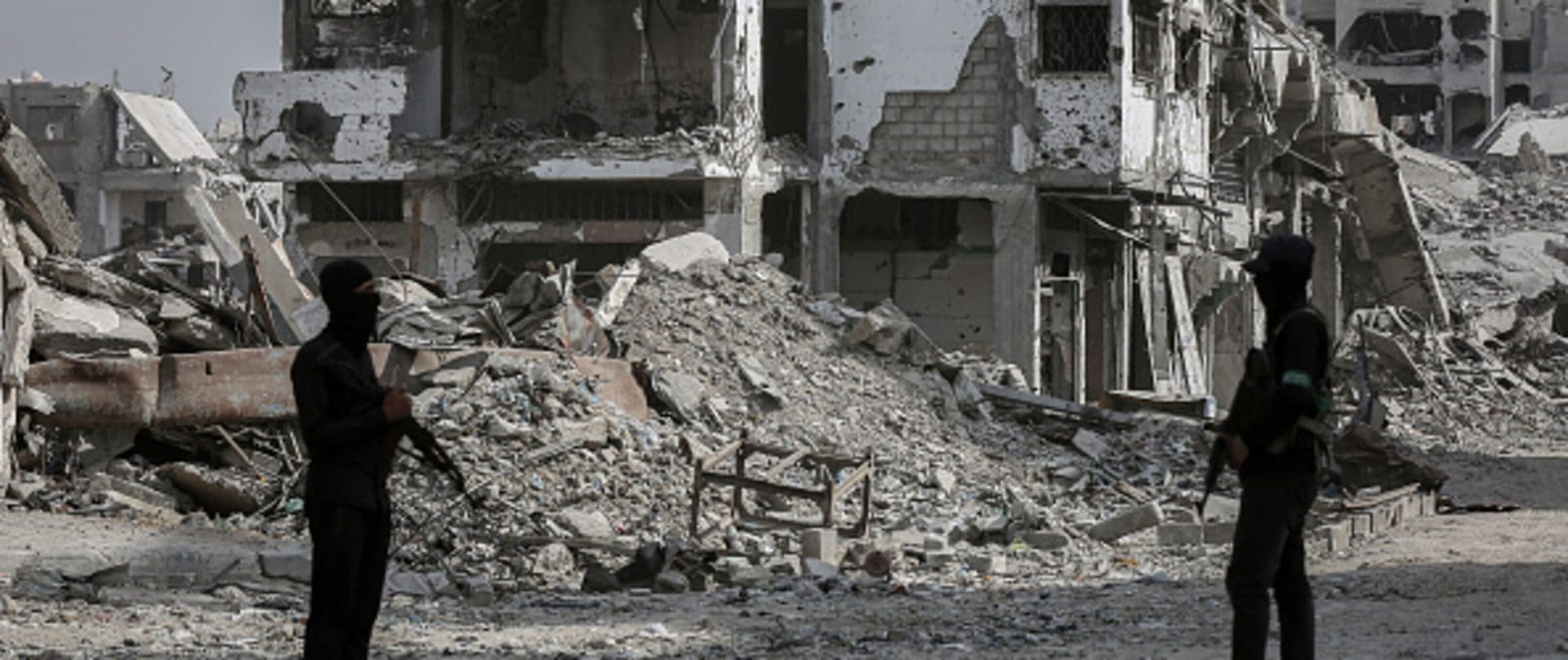 Two men wearing black with their faces covered holding rifles stand in front of the rubble of a destroyed building.