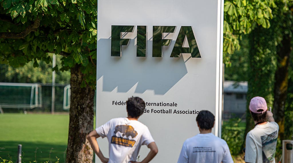 Three young people in tshirts look at a FIFA sign under leafy trees on a sunny day.