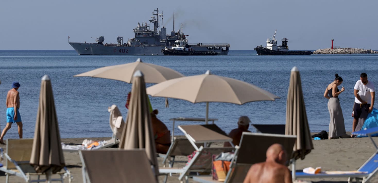 People on a beach with ships in the back.